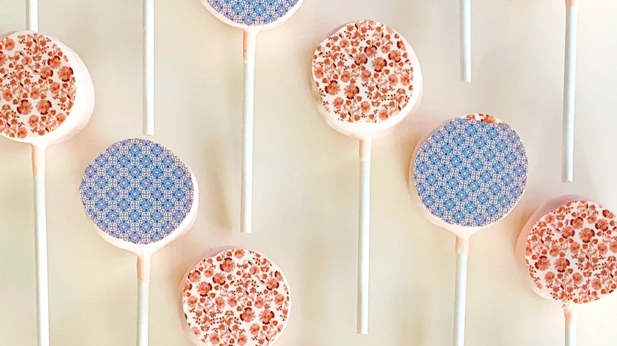 Close up image of customized cake pops with printed designs: one coral colored florals and one a blue "tile" pattern. Vanilla cake pops.